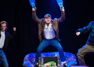 Szenenbild aus dem Theaterstück: Drei sich wild gebärdende Männer auf einer Theaterbühne, der mittlere steht auf einem Sofa und schwingt mit seinen Händen Schwimmflossen