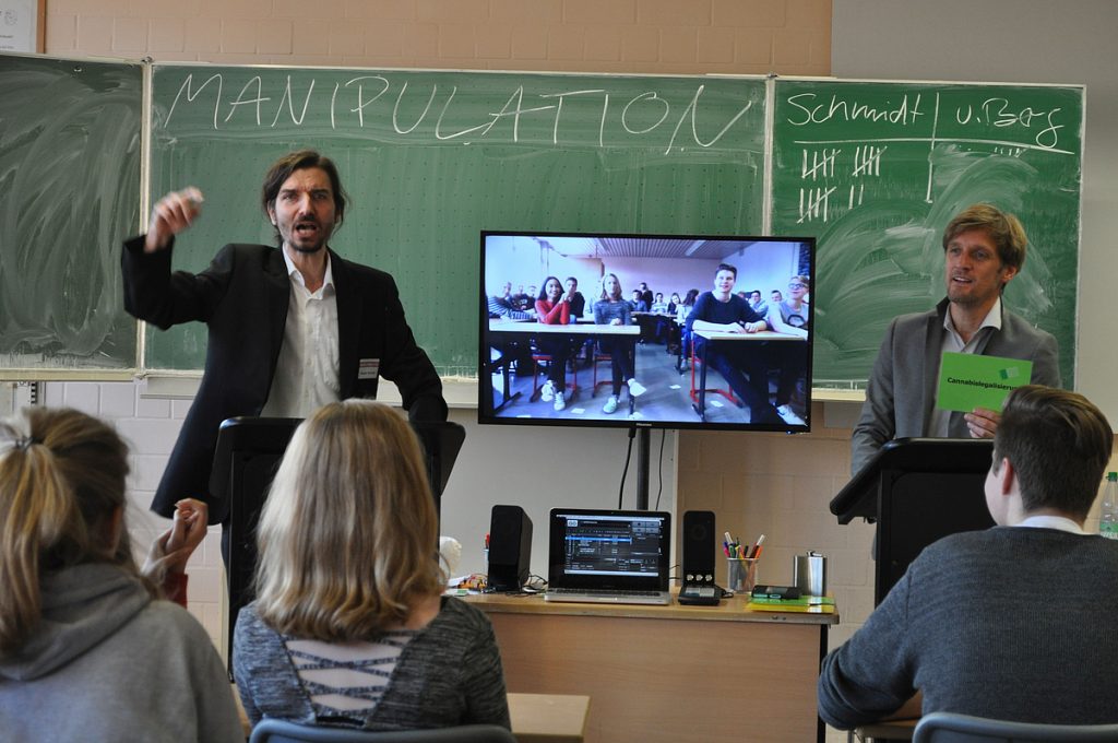 Zwei Personen stehen vor einer Klasse, einer gestikuliert. Auf der Tafel im Hintergrund steht das Wort “Manipulation”.
