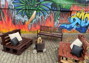 Drei selbstgebaute Sitzbänke gruppiert um einen Feuerkorb, im Hintergrund eine Wand mit Graffiti